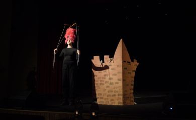 Na scenie osoba występująca ubrana na czarno. Trzyma rękoma za sznurki z marionetki. Obok niej dekoracja przedstawiająca mury zamku. Tło jest ciemne, widać na nim jedynie kilka drobnych światełek.