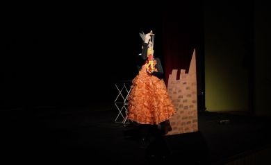 Na scenie aktorka w pomarańczowej sukni w ręku trzyma marionetkę. Obok dziewczyny stoi dekoracja jaką jest zamek. Ciemne tło. W oddali widać stojący na podłodze sceny stolik.