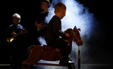 Trzej chłopcy. Jeden z nich siedzi na koniku na biegunach. Pozostali dwaj mają w rękach instrumenty przypominające gitarę. W tle widać ciężki dym.
