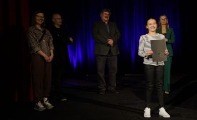 W tle dwie kobiety i dwóch mężczyzn. Z przodu dziewczyna w bluzce w biało-czarne paski z czarną teczka w ręku. Uśmiecha się.