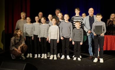 Na scenie grupka dzieci ubrana w koszulki w biało czarne paski. Jedne z chłopców w bluzce mającej wzór szachownicy trzyma czarną teczkę. Do zdjęcia wraz z dziećmi pozują dorośli: cztery kobiety i trzech mężczyzn.