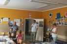 Na zdjęciu widać grupę osób w pomieszczeniu przypominającym salę lekcyjną lub świetlicę. W pomieszczeniu znajdują się: dwóch mężczyzn i kobieta, a także kilkoro dzieci. Jeden z mężczyzn gestykuluje, a kobieta trzyma w rękach książkę lub folder. Na ścianach wiszą plakaty, prace plastyczne i zdjęcia. Nad drzwiami znajduje się godło Polski. Na stolikach leżą arkusze papieru oraz różne przybory. W pomieszczeniu znajdują się stoły i krzesła.