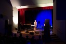 Na zdjęciu widać występ muzyczny na scenie, gdzie wokalistka śpiewa przy mikrofonie, a towarzyszą jej instrumentaliści grający na fortepianie i gitarze. Scena jest oświetlona nastrojowym, niebieskim światłem, a publiczność obserwuje koncert z widowni.