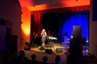 Na zdjęciu widać występ muzyczny na scenie, gdzie wokalistka śpiewa przy mikrofonie, a towarzyszy jej instrumentalista grający na fortepianie. Scena jest oświetlona nastrojowym, niebiesko-pomarańczowym światłem, a publiczność obserwuje koncert z widowni.