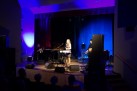 Na zdjęciu widać występ muzyczny na scenie, gdzie wokalistka śpiewa przy mikrofonie, a towarzyszą jej instrumentaliści grający na akordeonie i gitarze. Scena jest oświetlona nastrojowym, niebiesko-fioletowym światłem, a publiczność obserwuje koncert z widowni.