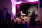 Na zdjęciu widać występ muzyczny na scenie, gdzie wokalistka śpiewa przy mikrofonie, a towarzyszy jej instrumentalista grający na fortepianie. Scena jest oświetlona nastrojowym, fioletowym światłem, a publiczność obserwuje koncert z widowni.