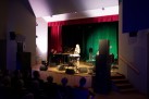 Na zdjęciu widać występ muzyczny na scenie, gdzie wokalistka śpiewa przy mikrofonie, a towarzyszą jej instrumentaliści grający na fortepianie i gitarze. Scena jest oświetlona nastrojowym, zielono-fioletowym światłem, a publiczność obserwuje koncert z widowni.