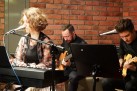 Kobieta przy keyboardzie. W tle dwaj mężczyźni grający na gitarach. Widać również ceglane ściany. 
