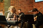 Kobieta przy keyboardzie. W tle dwaj mężczyźni grający na gitarach. Widać również ceglane ściany. 