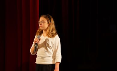 Dziewczynka w okularach, ubrana w białą bluzkę i czarną opaskę na głowie trzyma w prawej dłoni mikrofon. Patrzy przed siebie. 