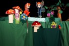 Na zdjęciu widać scenę teatralną z kolorowymi kukiełkami przypominającymi owady w bajkowej scenerii z grzybkami, kwiatami i drzewem. Dekoracje są wykonane z materiałów i papieru, tworząc fantazyjny, dziecięcy klimat.