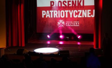Scena i widoczny na wyświetlaczu plakat dotyczący odbywającego się koncertu. Scenę odświetlają czerwone światła. 
