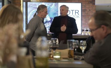 Dwóch mężczyzn jeden w okularach i szarej koszuli, drugi bez okularów w czarnej marynarce-rozmawiają. 