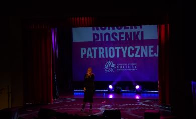Na scenie dziewczyna ubrana na czarno śpiewa do mikrofonu. W tle widać wyświetlany plakat dotyczący odbywającego się wydarzenia. 