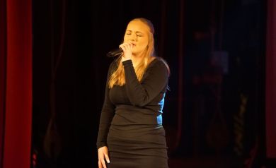 Dziewczyna o blond włosach, ubrana w czarną sukienkę śpiewa do mikrofonu. Ma przymknięte oczy. 