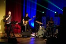 Na zdjęciu muzycy podczas swojego występu. Widać również lampy emitujące kolorowe światło. 