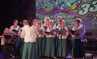 Zespół wokalny z dyrygentką, obok siedzi pani grająca na akordeonie. 