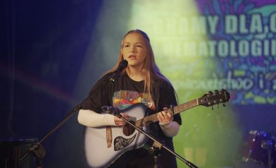 Dziewczyna z długimi, blond włosami grająca na gitarze i śpiewająca do mikrofonu. 