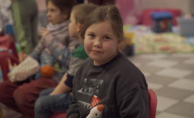 Dziewczynka z naklejonym na swetrze serduszkiem z Wielkiej Orkiestry Świątecznej Pomocy, uśmiecha się. W tle widać inne dzieci. 