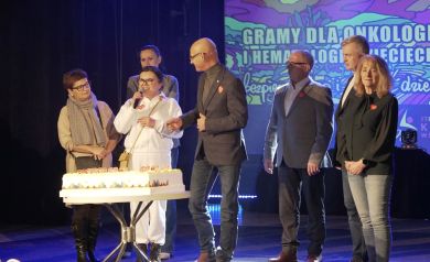 Kobiety i mężczyźni stojący na scenie. Kobieta ubrana na biało przemawia do mikrofonu. Przed nimi stoi tort. 