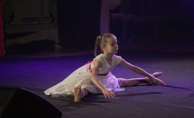 Dziewczynka w białej sukience z różowym paskiem wykonuje figurę taneczną. 