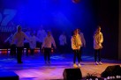Dzieci występujące na scenie. Wszystkie ubrane są w białe bluzy i ciemne spodnie. Scenę oświetla niebieskie i żółte światło, unosi się również tam ciężki dym. 