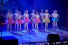 Dziewczynki w ubrane w białe bluzki i kolorowe, tiulowe spódniczki stoją na scenie.