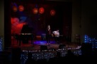 Scena ozdobiona jasnymi światełkami, na której występuje kobieta śpiewająca do mikrofonu i mężczyzna grający na gitarze. 