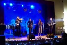 Na scenie zespół wokalno-instrumetalny w trakcie swojego występu. W zespole są zarówno mężczyźni jak i kobiety. 