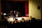 Na zdjęciu widać występ wokalistki wraz z sekcją instrumentalną (pianista, kontrabasista, perkusista). Wszyscy występujący muzycy są ubrani na czarno. Widać również część publiczności. 