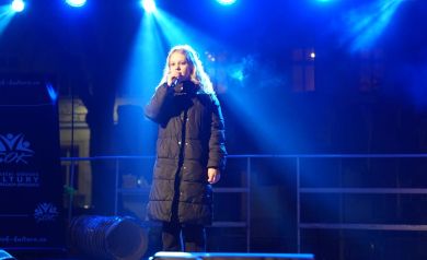 Dziewczyna występująca na scenie. Ubrana jest w czarną kurtkę. Scenę oświetla niebieskie światło. 