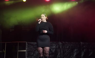 Dziewczyna w czarnej sukience stoi na scenie i śpiewa do mikrofonu. 