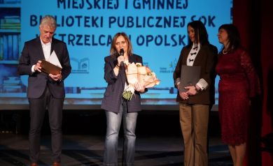 Na zdjęciu trzy kobiety i mężczyzna. Jedna z kobiet trzyma w dłoni bukiet i przemawia do mikrofonu.
