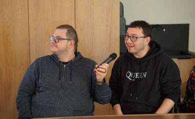 Dwóch mężczyzn ciemnych bluzach i okularach śpiewa do mikrofonu.