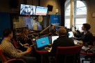 Zdjęcie przedstawia studio radiowe, Radio Opole, gdzie odbywa się nagranie lub audycja na żywo. W studiu znajduje się stół, przy którym siedzą cztery osoby. Osoby mają przed sobą mikrofony. Na stole widać laptopy, a także, słuchawki i kubek i inne przedmioty. W tle, za szklaną szybą, widoczne są dwa monitory oraz zegar. Na ścianie znajduje się logo „Radio Opole”.