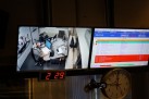 Zdjęcie przedstawia wnętrze studia radiowego, prawdopodobnie podczas audycji radiowej. Na ekranach widoczne są różne informacje, w tym: obraz z kamery, pokazujący dwie osoby siedzące przy biurku z mikrofonami i komputerami, zegar cyfrowy oraz tradycyjny zegar analogowy, ekran z harmonogramem audycji oraz liczniki czasu.
