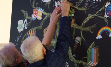 Mężczyzna i kobieta przyczepiają na czarnej tablicy, na której namalowane jest drzewo i jest napis ,,Drzewo Marzeń' znak graficzny. Mężczyzna ubrany jest w marynarkę w kratkę.