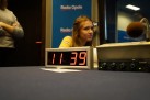 Zdjęcie przedstawia wnętrze studia radiowego, Radia Opole, na co wskazuje logo widoczne w tle. Na pierwszym planie znajduje się cyfrowy zegar, który wskazuje godzinę 11:39. Obok zegara widać konsolę mikserską oraz mikrofon. W kadrze widoczna jest również kobieta siedząca za stołem.
