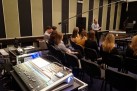 Zdjęcie przedstawia wykład lub warsztaty w Radiu Opole. Na pierwszym planie widać duży stół mikserski, a w głębi sali prelegent prowadzi zajęcia dla grupy słuchaczy.