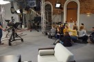 Na zdjęciu widać studio telewizyjne lub nagraniowe, w którym grupa młodych ludzi siedzi i rozmawia, podczas warsztatów. W pomieszczeniu znajduje się profesjonalny sprzęt filmowy, w tym kamery na statywach, lampy studyjne i inne elementy oświetlenia. Młodzi ludzie siedzą na podłodze i na kanapie, a jedna z osób gestykuluje, co może sugerować ożywioną dyskusję. W tle widać ceglaną ścianę, schody z metalową balustradą i kanapę.