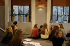 Zdjęcie przedstawia grupę młodych ludzi, prawdopodobnie uczniów, zgromadzonych w przestronnym pomieszczeniu z ceglanymi ścianami i dużymi oknami. Na parapecie stoją doniczki z roślinami. Siedzą na podłodze, rozmawiają między sobą. Obok nich leżą arkusze papieru. 