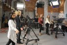 Zdjęcie przedstawia grupę osób w studiu telewizyjnym, wypełnionym różnym sprzętem, m. in. kamerami, czy lampami. 