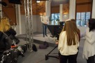 Zdjęcie ukazuje plan nagraniowy w studiu telewizyjnym. Na pierwszym planie widać sprzęt do nagrywania, w tym kamerę na statywie z zamontowanym teleprompterem. W tle stoi młody mężczyzna, prawdopodobnie prezenter lub aktor, natomiast w pobliżu kamery znajduje się ekipa nagraniowa, w tym kobiety z długimi włosami. W pomieszczeniu widoczne są też lampy oświetleniowe oraz ściany z cegły i duże okna.