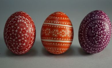 Na zdjęciu są trzy ozdobne jajka wielkanocne. Każde jest inaczej pomalowane i ozdobione białymi, precyzyjnymi wzorami kwiatowymi. Jajka stoją obok siebie na szarej powierzchni. Kolory jaj to czerwony, pomarańczowy i fioletowy. - powiększ
