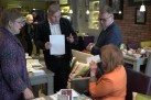 Zdjęcie przedstawia grupę czterech osób, w restauracji, podczas spotkania. Na pierwszym planie widać dwóch mężczyzn i dwie kobiety. Jeden z mężczyzn, ubrany w marynarkę, trzyma w ręku kartkę papieru i gestykuluje, jakby coś tłumaczył. Obok niego stoi drugi mężczyzna w okularach. Jedna z kobiet siedzi przy stole i trzyma kopertę przy twarzy, a druga stoi naprzeciwko mężczyzn, słuchając ich. Wnętrze lokalu ma ceglane ściany i jest oświetlone lampami wiszącymi. Na stołach widoczne są filiżanki z kawą i inne przedmioty. W tle widać inne osoby oraz elementy wystroju, takie jak obrazy na ścianach.