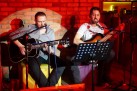 Dwóch mężczyzn grających na gitarach w oświetlonej czerwonym światłem restauracji. Jeden z nich śpiewa do mikrofonu. 