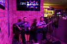 Dwóch mężczyzn grających na gitarach i śpiewających. W tle widać ekran wiszący na ceglastej ścianie i część baru. Całość oświetlona jest różowym światłem. 