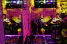 Dwóch mężczyzn grających na gitarach i śpiewających. Widać sprzęt muzyczny, ekran wiszący na ceglastej ścianie i część baru. Całość oświetlona jest żółto-różowym światłem. 