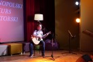 Na zdjęciu młody chłopak grający na gitarze prawdopodobnie śpiewa do mikrofonu znajdującego się na stojaku. 