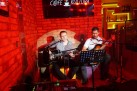 Dwóch mężczyzn grających na gitarach i śpiewających. Całość oświetlona jest czerwonym światłem. 